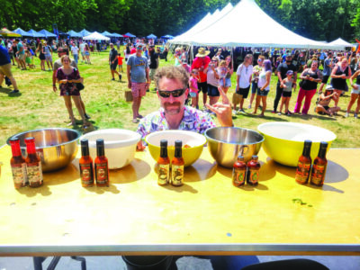man sitting behind table with rows of bowls and varieties of hot sauce in front of bowls, event tents and crowds of people in background, sunny day on grassy field