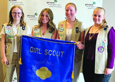 4 girls wearing girl scout vests standing in row and holding banner with the girl scout logo