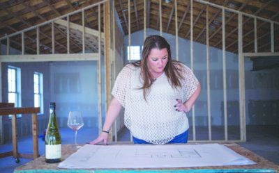 woman standing in room being renovated, looking at architectural drawing on table beside bottle of wine and wine glass