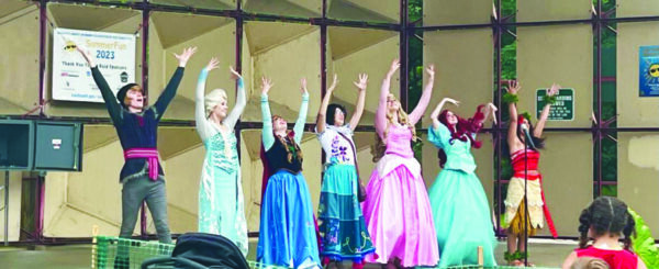 actors dressed as disney princess characters on stage with hands raised as they sing at outdoor venue