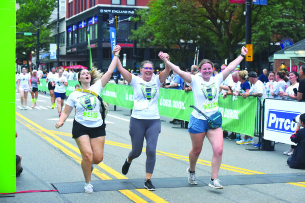 3 women running beside each other, holding raised up hands as they celebrate  during road race, people watching from sidewalk