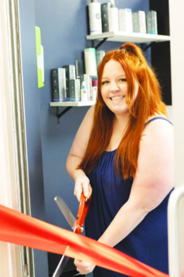 young woman with long hair, smiling, cutting ceremonial ribbon in front of shelf containing beauty products