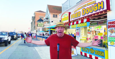 man wearing straw boater hat, arms raised in wide gesture, standing in front of microphone on sidewalk, fried dough stand behind him, beach town