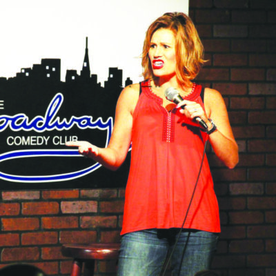 woman holding microphone on stage in front of comedy club sign, making weird facial expression