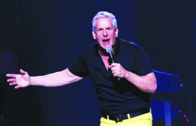 man with white hair, holding microphone, gesturing to the side with outstretched hand as he speaks on stage