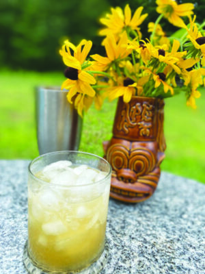 short glass filled with cloudy cocktail and ice, on table outside beside decorative vase filled with sunflowers