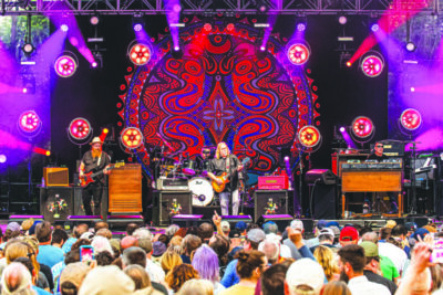 band members on stage in front of crowd, outdoor venue, colorful mandala on back wall of stage