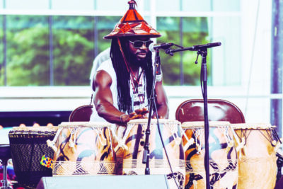 male musician outside playing large drums with his hands, wearing conical hat, dreadlocks and sunglasses, sunny day