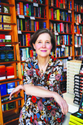 mature woman posing in bookstore in front of wall of wooden shelves, beside table with piles of books