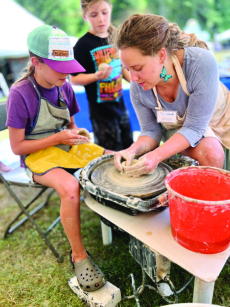woman teaching 2 children how to make clay pot on wheel at outdoor demonstration