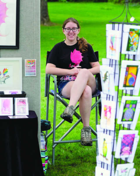 young woman sitting on outdoor folding chair behind racks of printed artwork on green lawn
