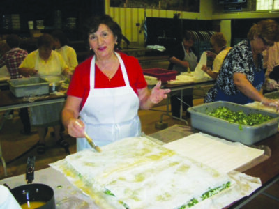 woman in large room at community center, assembling large sheet of spanakopita with other volunteers