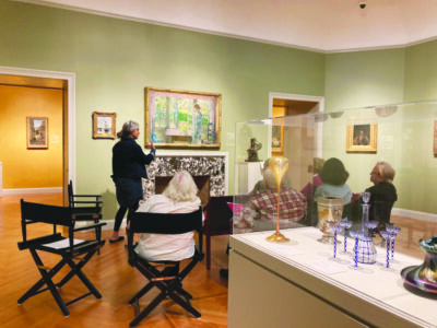 in art gallery, woman standing and pointing to painting while speaking to group of seated elderly people