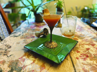 cocktail with marshmallow garnish in martini glass with decorative stem, sitting on square plate on long table