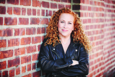 woman with curly red hair standing in front of brick building, arms folded, wearing leather coat, smiling