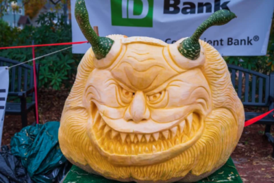 giant pumpkin sculpturally carved into pointy-toothed leering face with gourds making horns at top of head