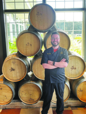 man standing in front of wine barrels, wearing chef's jacket, smiling