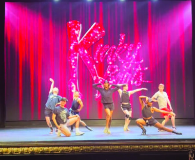 group of diverse male dancers posing on stage in front of red curtain with sparkly words Kinky Boots hanging