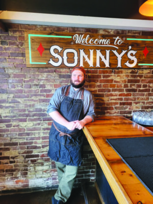 bearded man wearing chef's apron and backward baseball cap, leaning against counter, back to brick wall, under signs that reads "Welcome to Sonny's"