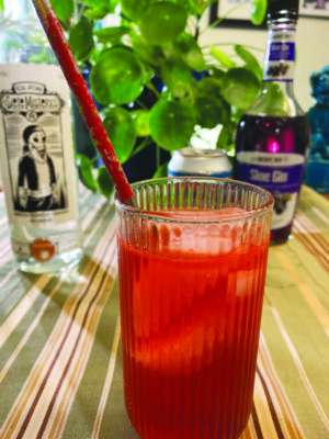reddish cocktail with ice in tall ridged glass, with straw, on striped table cloth, bottle and plant in background