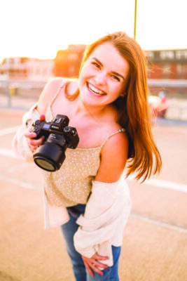 young woman holding camera, smiling and posing outside