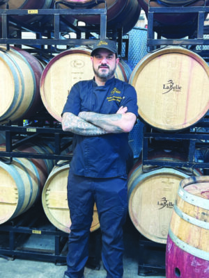 man with beard, wearing Labelle Winery baseball cap and black chef's jacket, standing in front of barrels of wine, arms crossed