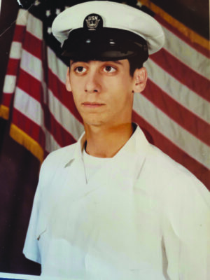 young man wearing white uniform with Navy hat, standing in front of American flag