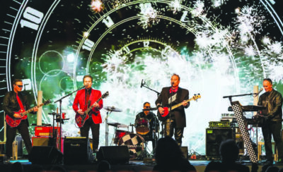 5 men playing instruments on stage in front of screen showing old fashioned clock face with small fireworks