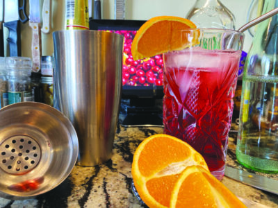 cranberry cocktail in tall cut glass, with orange garnish and metal straw, on countertop beside cocktail shaker