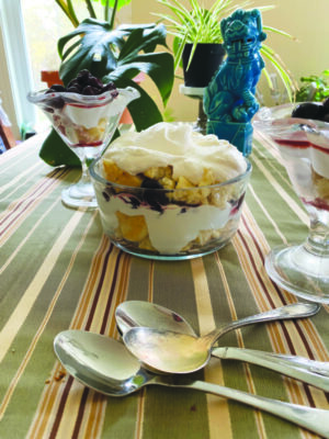 glass bowl filled with layers of broken cookies and whipped cream, on talbe with striped table cloth, spoons lying beside