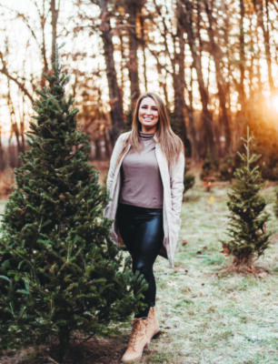 woman wearing open sweater at sunset on fall day, standing near small pine tree, woods behind