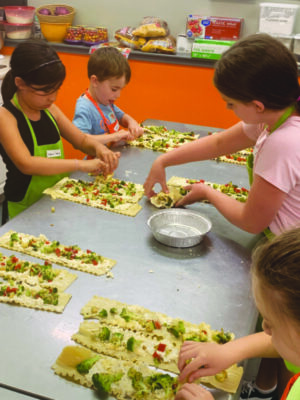 young children gathered around table in cooking lesson, rolling vegetables into strips of dough