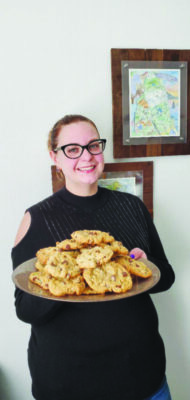 woman wearing glasses standing in front of wall with photos, holding up large plate piled with cookies