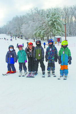 children dressed up in snow pants and coats, standing in a line on ski slope with skis on snowy day