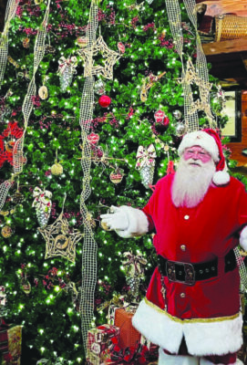 man in santa suit standing in front of very tall christmas tree decorated with large glass ornaments