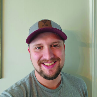 Man with some scruff smiling at the camera wearing a baseball cap and grey t-shirt