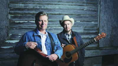 2 middle aged men wearing button up shirts and jackets, holding guitars, standing in front of wooden wall with peeling paint