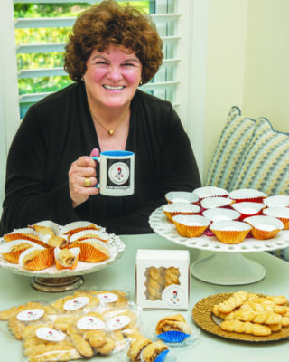 curly haired middle aged woman sitting behind table displaying greek pastry on plates and some wrapped for sale, holding mug, smiling