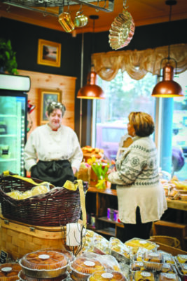 woman wearing apron standing in store filled with baked goods talking with another woman, cozy scene at dusk