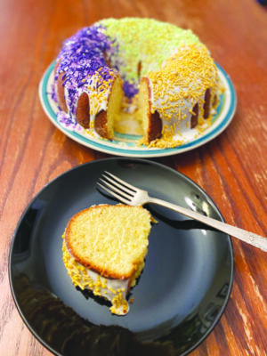 round cake with hole in the middle, covered in icing, decorated with yellow, green and purple sprinkles, one piece cut out and set on plate beside