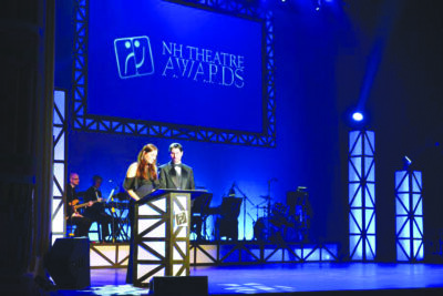 woman and man standing behind podium , on darkened stage, orchestra behind them, screen with words NH Theatre Awards