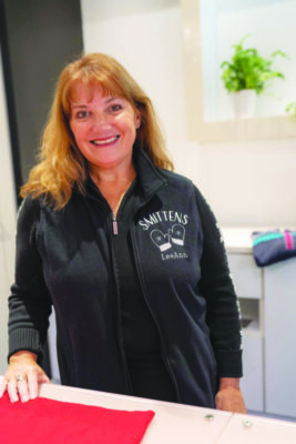 woman standing behind table in sewing workroom, wearing black jacket with Smittens Mittens logo on left chest, smiling