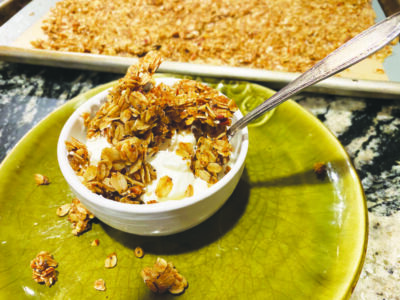 small bowl of granola with milk, spoon sticking out, bowl sitting on plate beside sheet pan containing a flat layer of baked granola