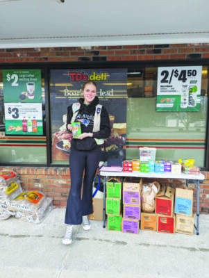 girl scout standing next to table filled with cookies outside of storefront