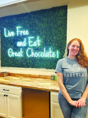 Woman standing in front of illuminated sign that reads "Live Free and Eat Great Chocolate!"