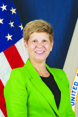 Woman with short blonde hair smiling at camera wearing bright green blazer with the American flag in the background