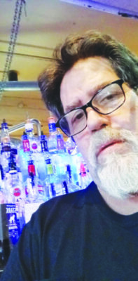 headshot of man with beard, wearing glasses, leaning into the photo with bottles of alcohol on the shelves behind him