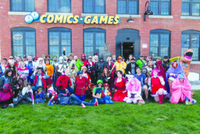 large group of people posing on lawn in front of brick building with large windows, sign above reads Comics/Games. Everyone dressed in cosplay.