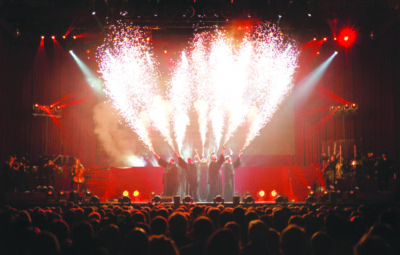 singers dressed in black robes standing in middle of stage, red colored lights and pyrotechnics behind them