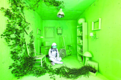 person dressed in astronaut suit, sitting in room painted all green with green chairs, empty picture frames and other things, fake vines coming out of box and crawling up wall